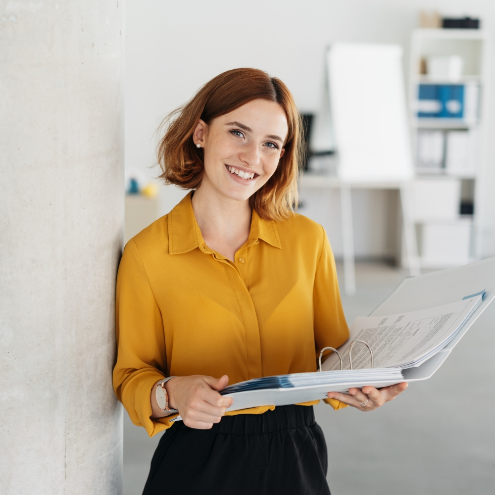 Hymyilevä punahiuksinen nainen nojaa toimiston seinään papereita käsissään.