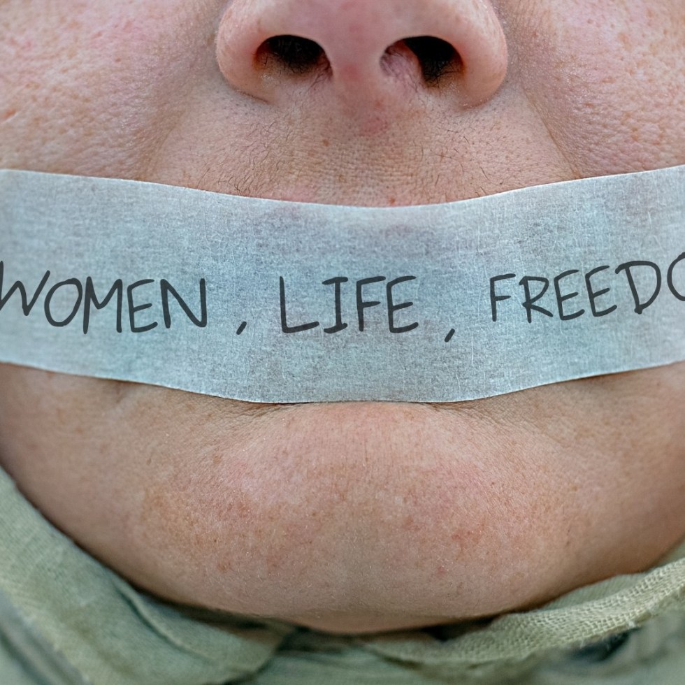 Kuva naisen kasvojen alaosasta. Suu on peitetty teipillä, jossa lukee Women, Life, Freedom.