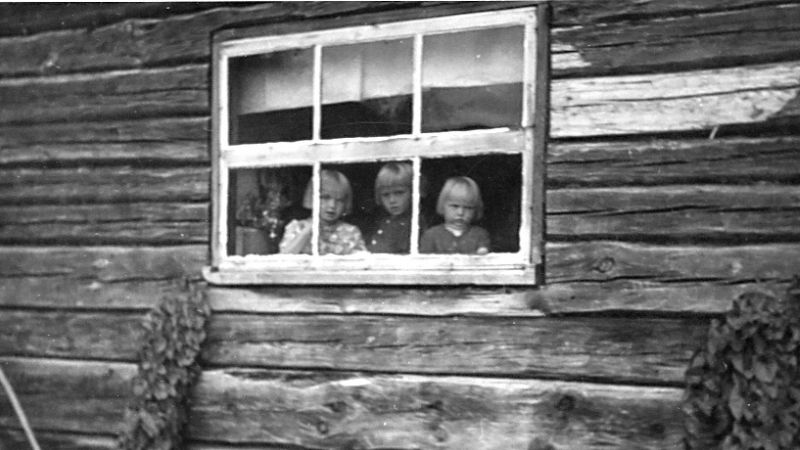 Kolme lasta kurkistaa pirtin ikkunasta ulos.