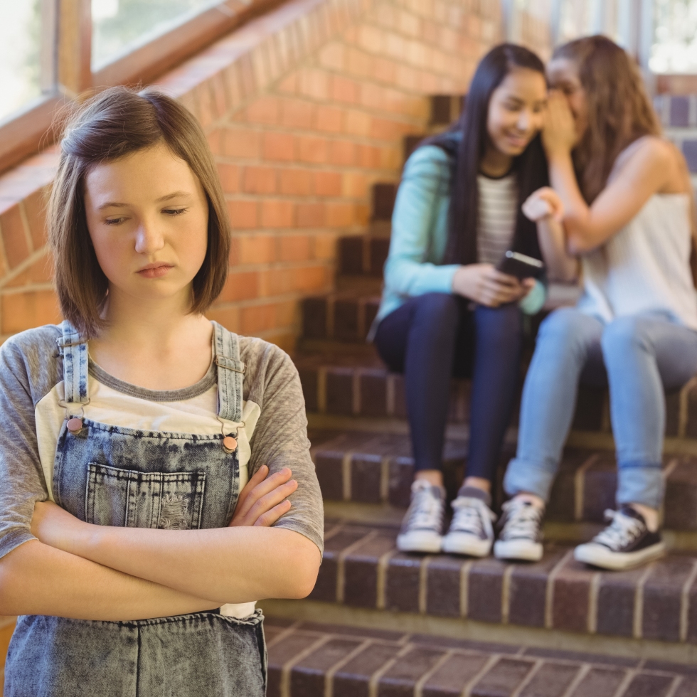 Kuva koulun portaiskosta, kaksi tyttöä istuu supisemassä taustalla ja etualalla yksinäinen tyttö katselee alaspäin murheissaan.